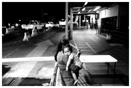 Fotografia em preto e branco de uma mulher de costas segurando uma criança no colo junto a uma mureta em um posto de fronteira; ao fundo, pista com veículos e passarela coberta.