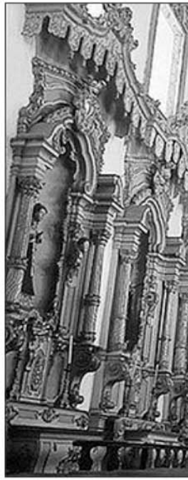 Fotografia em preto e branco do interior da igreja, destacando o altar ricamente decorado com talha dourada e estátuas religiosas.
