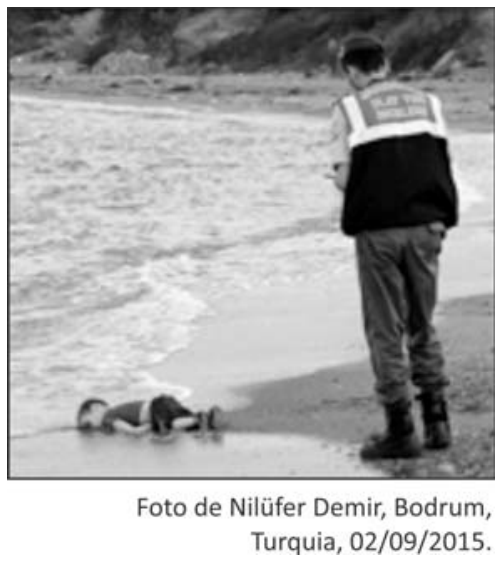 Fotografia em preto e branco mostrando um agente oficial de costas observando o corpo de uma criança (Alan Kurdi) deitada de bruços na areia da praia, onde quebram as ondas. Abaixo, legenda: Foto de Nilüfer Demir, Bodrum, Turquia, 02/09/2015.