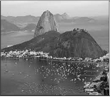 Fotografia do Pão de Açúcar e de morros próximos, com encostas parcialmente cobertas por vegetação, construções junto à costa e numerosos barcos na baía em primeiro plano.