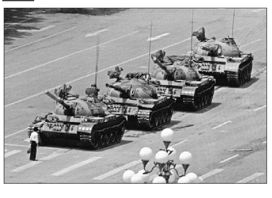 F1 Fotografia em preto e branco mostrando uma fileira de quatro tanques de guerra em uma ampla avenida em Beijing, China. À frente do primeiro tanque, à esquerda, uma pessoa solitária está de pé no meio da via, bloqueando a passagem. No primeiro plano inferior, vê-se o topo de um poste com luminárias esféricas brancas.