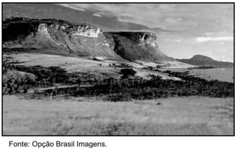 Fotografia em preto e branco de uma paisagem de chapada, com escarpas íngremes e topo plano ao fundo, e vegetação rasteira e arbustiva em primeiro plano. Abaixo da imagem lê-se: "Fonte: Opção Brasil Imagens."