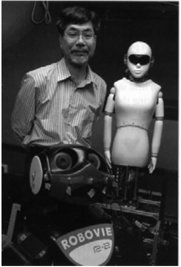Fotografia de um pesquisador em pé ao lado de um robô humanoide e, em primeiro plano, outro robô com a inscrição "ROBOVIE R-2".