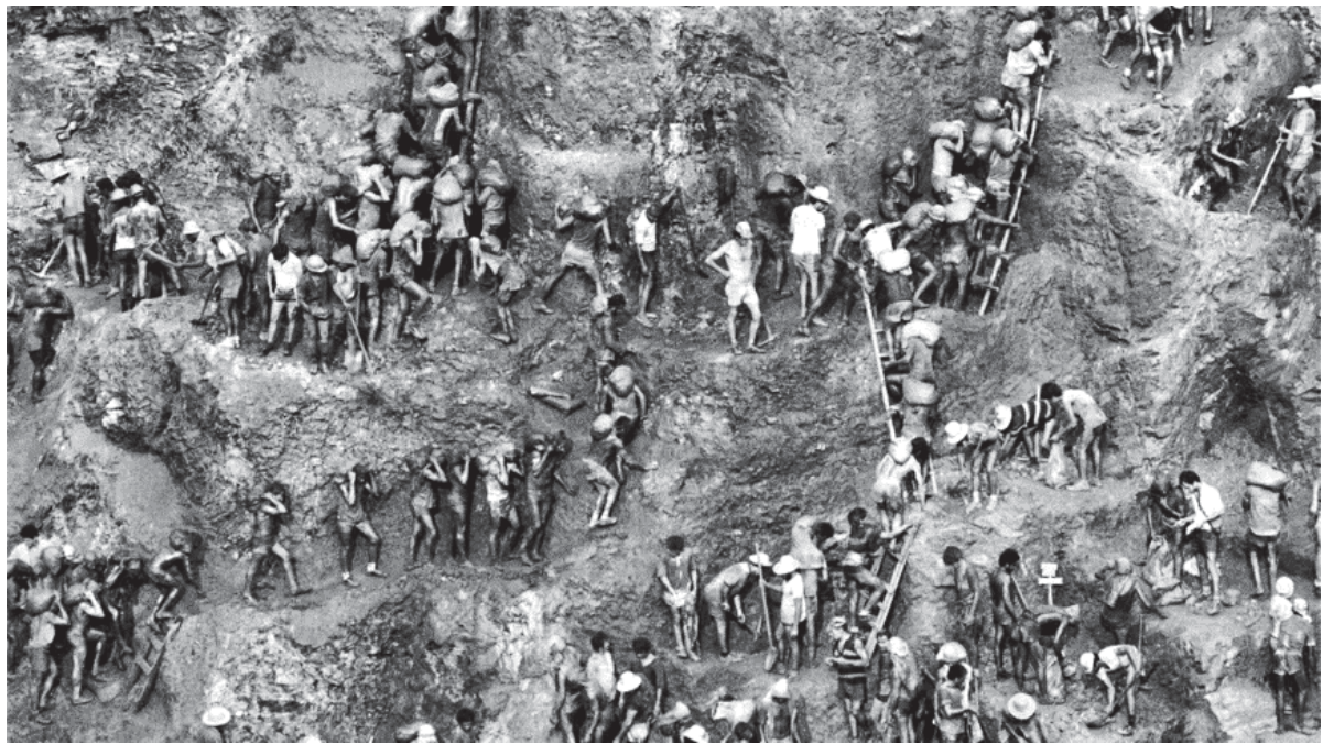 Fotografia em preto e branco de Serra Pelada, mostrando numerosos garimpeiros espalhados por encostas e patamares de terra de um garimpo a céu aberto. Muitos carregam grandes sacos sobre os ombros ou a cabeça, enquanto outros sobem e descem escadas de madeira apoiadas nos barrancos.