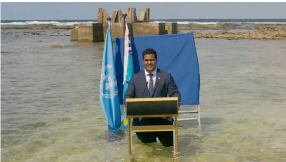 Simon Kofe discursando em um púlpito dentro do mar com água até os joelhos, com bandeiras ao fundo.