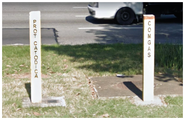 Fotografia em plano médio de dois marcos verticais de concreto (totens) fincados em uma calçada gramada ao lado de uma via urbana. O totem à esquerda traz a inscrição vertical 'PROT CATÓDICA' e o da direita traz a inscrição vertical 'COMGAS'.