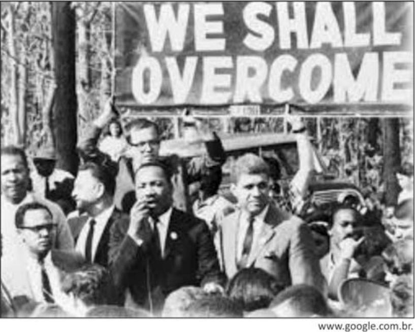 Fotografia em preto e branco de uma manifestação, com Martin Luther King em destaque entre outros participantes. Acima deles, uma faixa traz a inscrição “WE SHALL OVERCOME”. No canto inferior direito, consta o crédito “www.google.com.br.”.