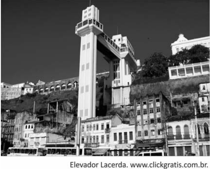 Fotografia em preto e branco do Elevador Lacerda, em Salvador, com suas torres e a passagem elevada entre elas, ligando a parte baixa da cidade à parte alta. Ao redor, aparecem edifícios junto ao desnível do terreno e veículos na rua inferior. Legenda: “Elevador Lacerda. www.clickgratis.com.br.”