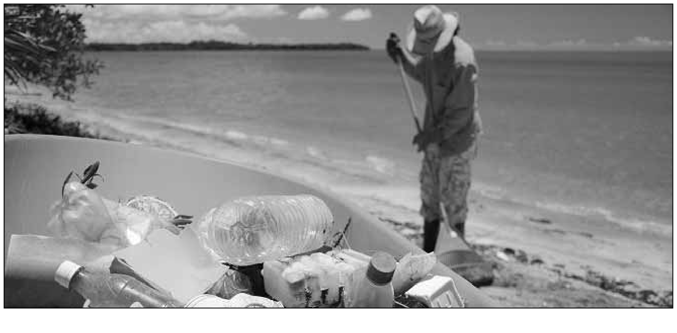 Fotografia em preto e branco mostrando resíduos plásticos acumulados em primeiro plano, como garrafas e potes, e ao fundo uma pessoa com chapéu rastelando a areia de uma praia à beira-mar.