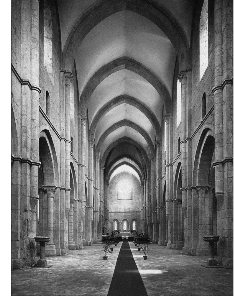 Fotografia em preto e branco do interior da Abadia de Fossanova, mostrando uma nave longa e alta, ladeada por pilares de pedra e arcos ogivais, com uma sequência de abóbadas de arcos apontados no teto. Janelas altas iluminam o espaço, e um tapete escuro percorre o corredor central até o altar ao fundo.