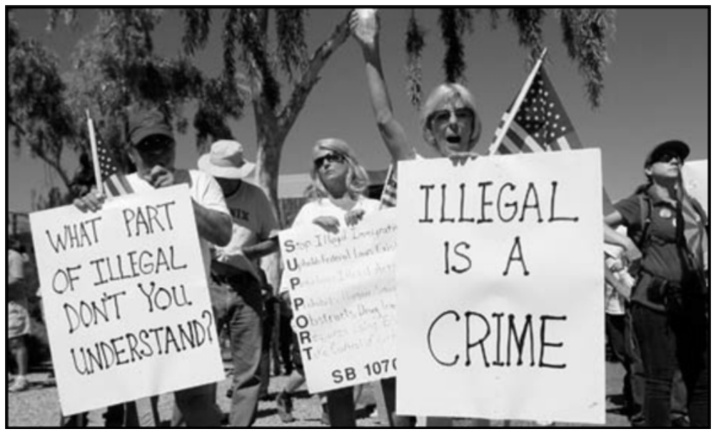 Manifestantes segurando cartazes em protesto contra a imigração ilegal nos Estados Unidos, com dizeres como "WHAT PART OF ILLEGAL DON'T YOU UNDERSTAND?" e "ILLEGAL IS A CRIME".