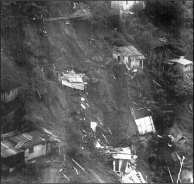 Fotografia em preto e branco de uma encosta íngreme ocupada por pequenas casas. Uma ampla faixa de solo exposto atravessa a encosta, com destroços de construções e casas atingidas pelo escorregamento; há vegetação e outras casas nas laterais.