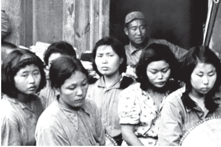 F2 Fotografia em preto e branco de um grupo de mulheres asiáticas, algumas olhando para baixo, reunidas diante de uma construção, com um homem de boné ao fundo.