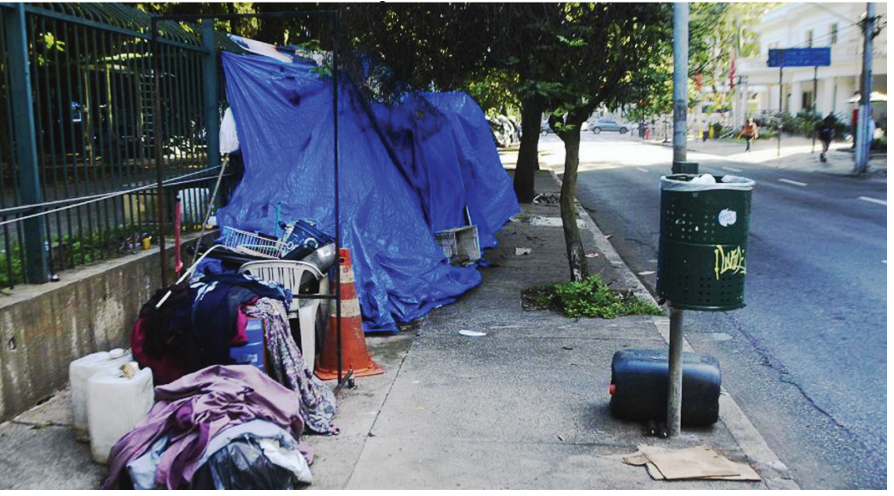 Fotografia de calçada em São Paulo mostrando barracas improvisadas cobertas com lonas azuis montadas rente a uma grade, com pertences acumulados como roupas, galões plásticos e cadeiras, ao lado de uma lixeira de rua fixada em poste metálico.