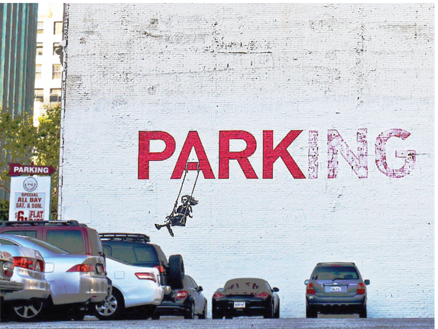 F1 Obra de arte urbana de Banksy em uma parede de tijolos brancos de um estacionamento com carros parados à frente. Na parede está pintada em vermelho a palavra "PARKING", com as letras "ING" desbotadas, destacando a palavra "PARK"; preso à letra "A", há o desenho de uma menina em um balanço.