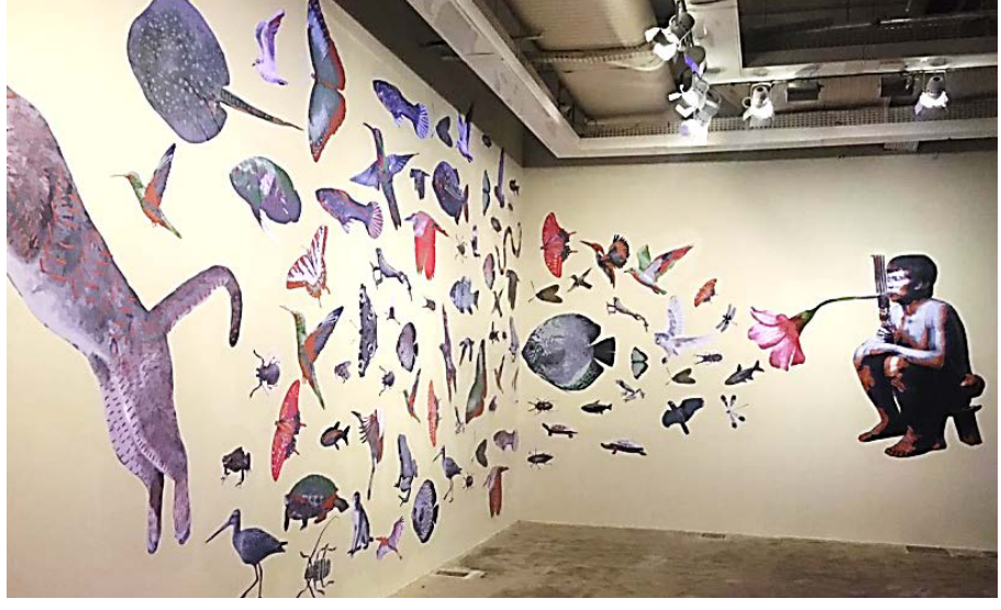 Fotografia da instalação artística de Denilson Baniwa em uma sala de exposição, com a técnica de lambe-lambe sobre paredes claras. À direita, vê-se a figura de uma pessoa indígena agachada, com adorno na cabeça e assoprando através de um instrumento de sopro em direção a uma flor, de onde se projeta uma profusão de gravuras de animais silvestres (peixes, aves, borboletas, insetos, tartaruga, arraia e um grande felino à esquerda) que se espalham cobrindo duas paredes contíguas.