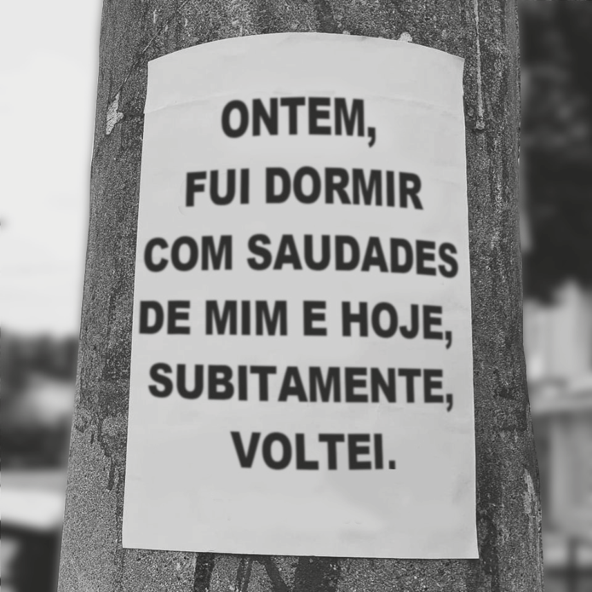 Fotografia de um cartaz lambe-lambe colado em um poste de concreto, com a frase escrita em letras maiúsculas: "ONTEM, FUI DORMIR COM SAUDADES DE MIM E HOJE, SUBITAMENTE, VOLTEI."
