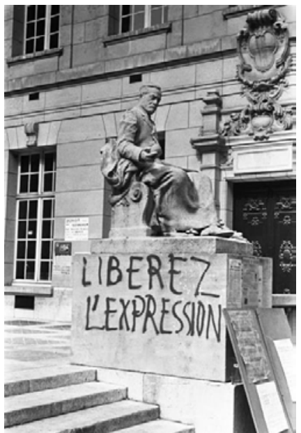 Fotografia em preto e branco de uma estátua sentada sobre uma base de pedra em frente a um prédio em Paris. Na base da estátua, está pichada em letras maiúsculas a frase "LIBEREZ L'EXPRESSION".
