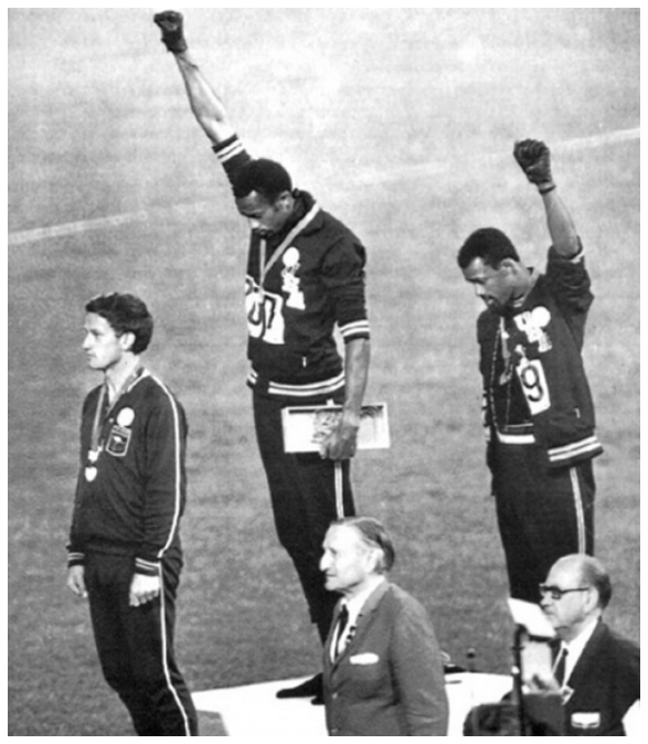 Fotografia em preto e branco do pódio dos 200 metros rasos nos Jogos Olímpicos de 1968, no México. No centro, no degrau mais alto, o atleta Tommie Smith está de cabeça baixa com o punho direito enluvado de preto erguido para o alto. À sua direita (à esquerda na imagem), no segundo lugar, está o atleta australiano Peter Norman, e à sua esquerda (à direita na imagem), no terceiro lugar, está John Carlos, também de cabeça baixa e com o punho esquerdo enluvado erguido para o alto.