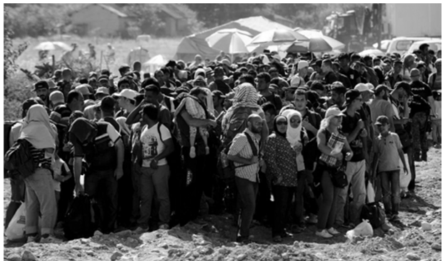 F1 Fotografia em preto e branco mostrando uma grande multidão de imigrantes e refugiados aglomerados ao ar livre, muitos carregando mochilas e sacolas, com algumas tendas e policiais ao fundo.