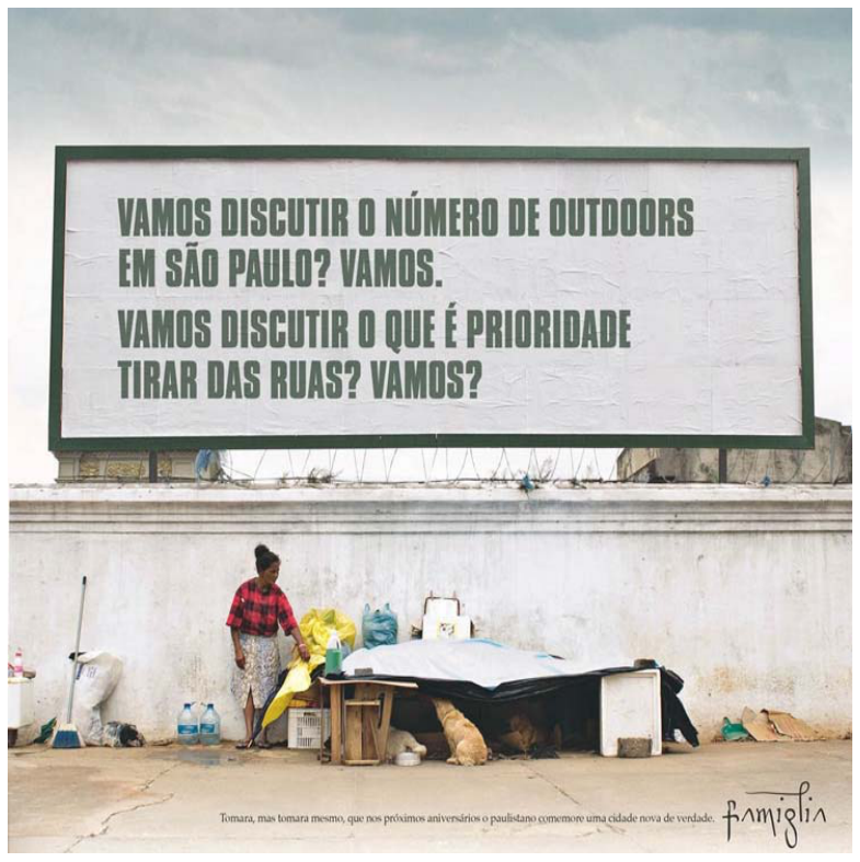 Anúncio publicitário com a fotografia de um grande outdoor posicionado acima de um muro. No outdoor está escrito em letras garrafais verdes: "VAMOS DISCUTIR O NÚMERO DE OUTDOORS EM SÃO PAULO? VAMOS. VAMOS DISCUTIR O QUE É PRIORIDADE TIRAR DAS RUAS? VAMOS?". Na calçada em frente ao muro, há uma mulher em situação de rua ao lado de um abrigo improvisado de lonas e móveis velhos, acompanhada de cachorros. No canto inferior da imagem, em letras pequenas, lê-se uma frase e a assinatura da agência Famiglia.