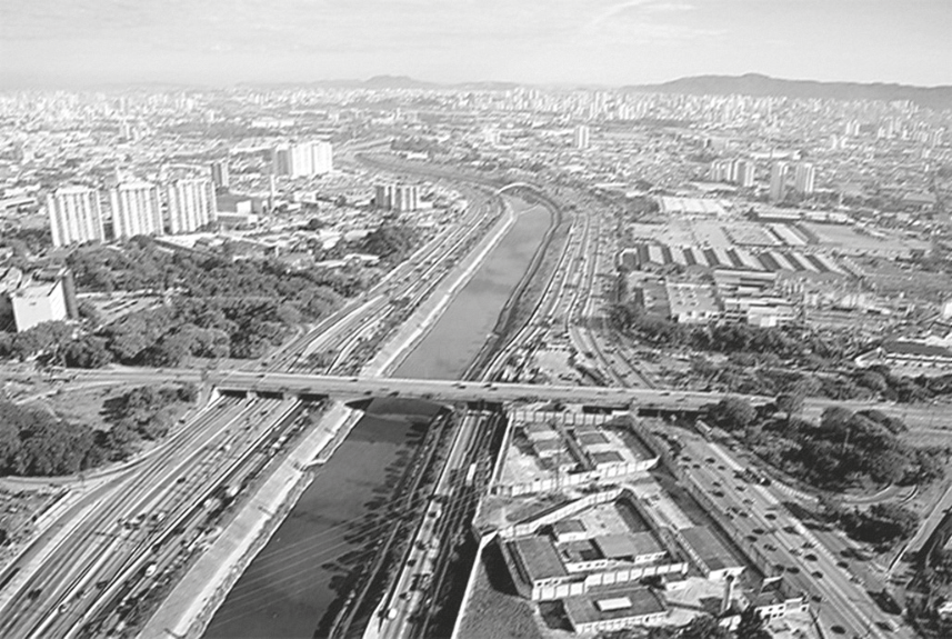 Fotografia aérea do Rio Tietê, em São Paulo, retificado e canalizado, margeado por pistas de tráfego expressas (Marginal Tietê), com um viaduto transversal e densa área urbana ao redor, com prédios e construções industriais.