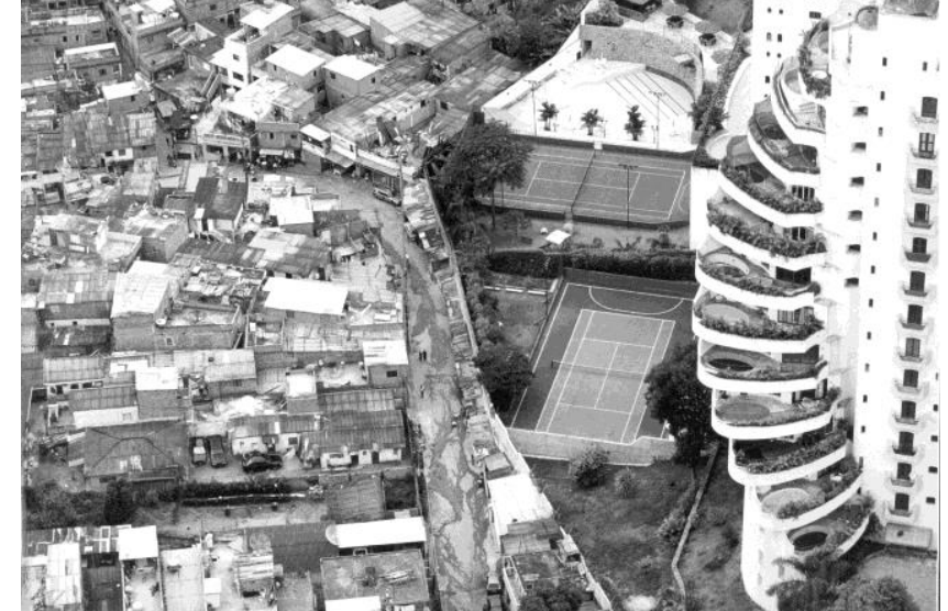 Fotografia aérea exibindo a divisa física entre a favela de Paraisópolis, à esquerda, densamente ocupada por habitações precárias e estreitas vielas, e um condomínio residencial de luxo no Morumbi, à direita, com prédio de alto padrão com piscinas nas varandas, quadras de tênis e piscinas privativas, separados por um muro.