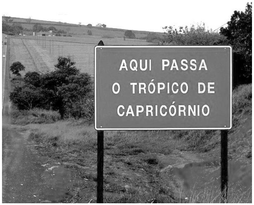 F1 Fotografia em preto e branco com uma placa retangular em primeiro plano onde está escrito: "AQUI PASSA O TRÓPICO DE CAPRICÓRNIO". Ao fundo, observa-se uma paisagem rural com vegetação, uma estrada à esquerda e colinas ao longe.