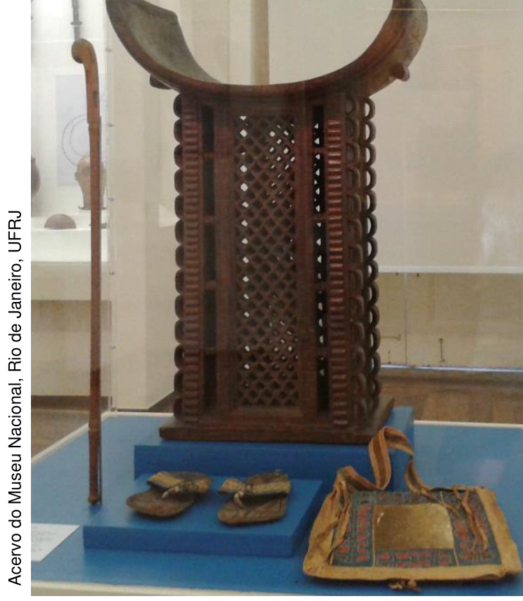 Fotografia em vitrine de museu mostrando o trono do rei Adandozan, esculpido em madeira com assento côncavo, ao lado de uma bengala à esquerda, e à frente um par de sandálias e uma bolsa cerimonial.