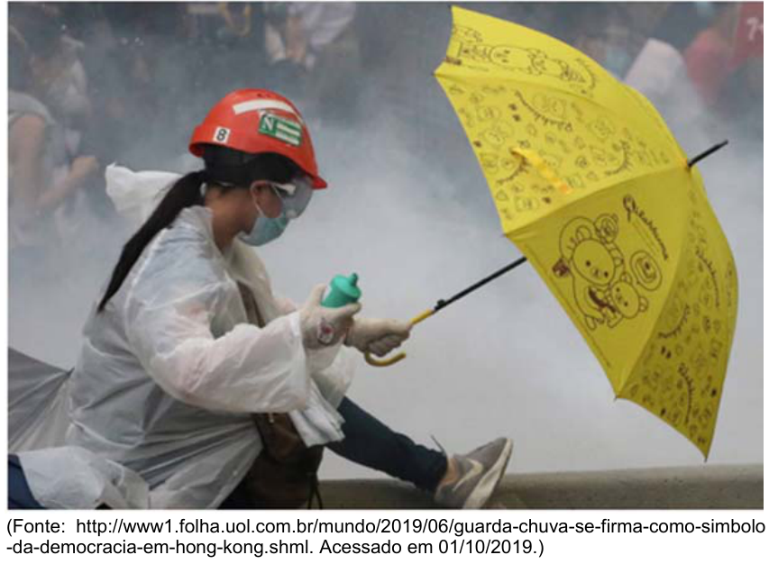 Manifestante em Hong Kong usando capacete laranja, capa de chuva, óculos de proteção e máscara facial, agachado e segurando um guarda-chuva amarelo em meio a fumaça de gás.