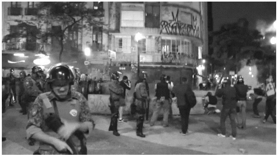 Fotografia em preto e branco mostrando policiais militares equipados atuando durante a noite em frente a um edifício urbano na reintegração de posse de um imóvel ocupado.