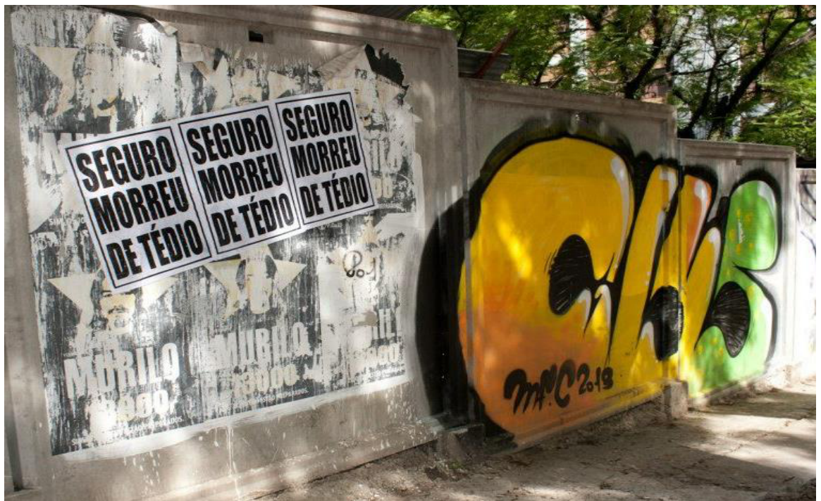 Fotografia de uma intervenção urbana em um muro de concreto, onde estão colados três cartazes idênticos com a inscrição "SEGURO MORREU DE TÉDIO" em letras maiúsculas garrafais pretas sobre fundo branco. Ao lado dos cartazes, há um grafite colorido com formas orgânicas nas cores amarela e verde.