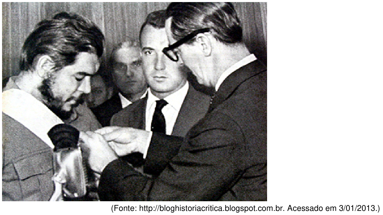 Fotografia em preto e branco na qual o presidente Jânio Quadros, visto de perfil à direita com óculos e paletó escuro, condecora Ernesto Che Guevara, à esquerda, com a Ordem Nacional do Cruzeiro do Sul. Ao fundo, homens de terno observam a cena. Abaixo da imagem lê-se: (Fonte: http://bloghistoriacritica.blogspot.com.br. Acessado em 3/01/2013.).