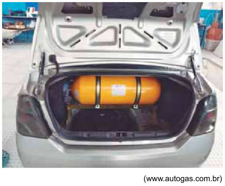 Fotografia do porta-malas aberto de um carro prateado, com um cilindro amarelo de GNV instalado horizontalmente e preso por cintas metálicas.