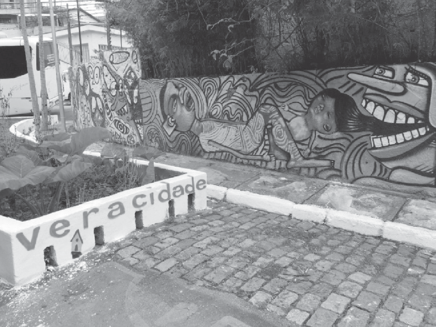 Fotografia de um espaço urbano com rua de paralelepípedos e muros grafitados. Em primeiro plano, uma mureta branca traz escrita a palavra "veracidade", com o "A" estilizado em formato de casa. Ao fundo, um muro exibe grafites com figuras humanas expressivas e linhas sinuosas.