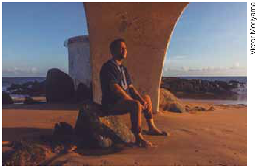 Foto de Itamar Vieira Junior sentado em uma rocha sob uma estrutura de alvenaria em arco na praia, olhando para o mar ao entardecer.