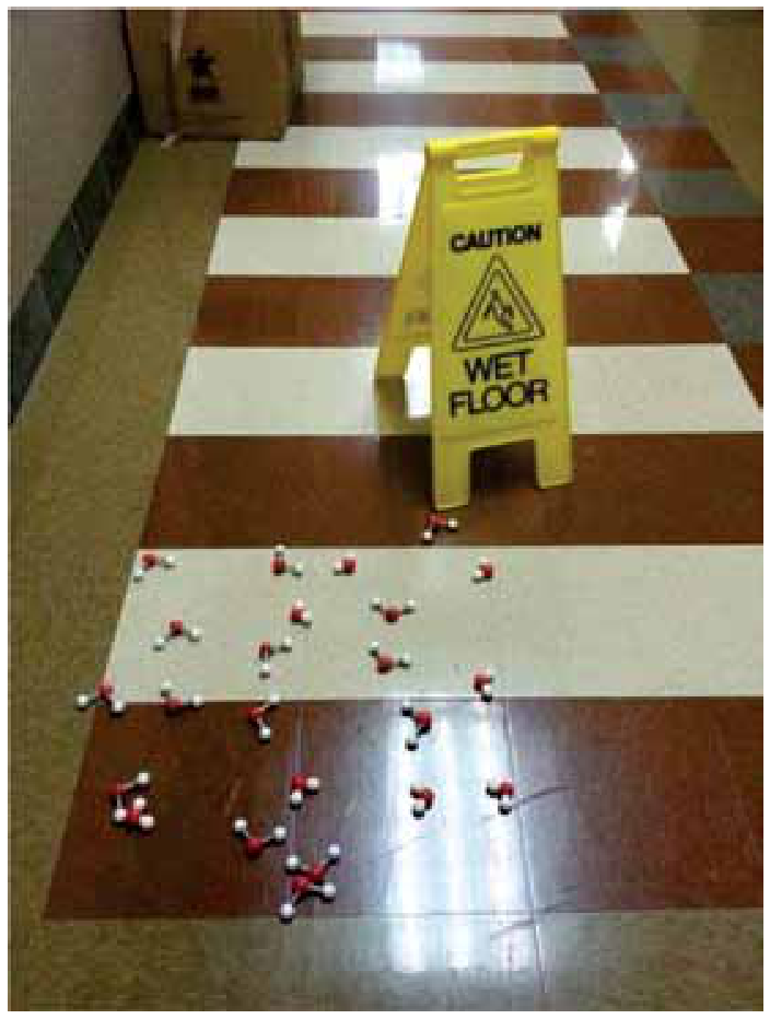 Fotografia de um corredor com uma placa amarela de alerta onde se lê "CAUTION WET FLOOR" e, espalhados pelo chão à sua frente, diversos modelos moleculares de bolas e varetas representando moléculas de água (átomos vermelhos de oxigênio ligados a dois átomos brancos de hidrogênio).