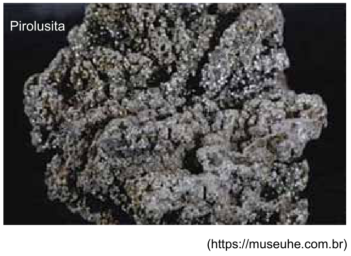 Fotografia em plano aproximado de uma amostra do mineral pirolusita sobre fundo escuro. O mineral apresenta aspecto rugoso, poroso e coloração cinza metálica prateada com cavidades escuras. No canto superior esquerdo, lê-se "Pirolusita".