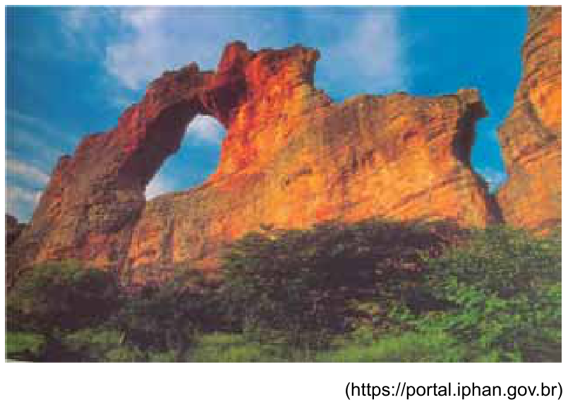 Fotografia da Pedra Furada, no Parque Nacional da Serra da Capivara, destacando um grande arco rochoso de tons avermelhados cercado por vegetação e com o céu ao fundo. Abaixo, à direita, lê-se: (https://portal.iphan.gov.br)