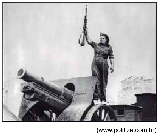 Fotografia em preto e branco retratando uma mulher miliciana em pé sobre um canhão de artilharia militar, erguendo um fuzil verticalmente com a mão direita.