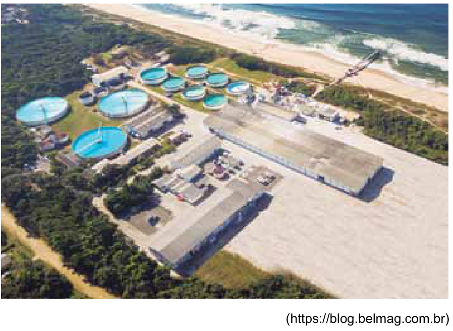 Vista aérea de uma instalação industrial próxima à praia com diversos tanques circulares e galpões.