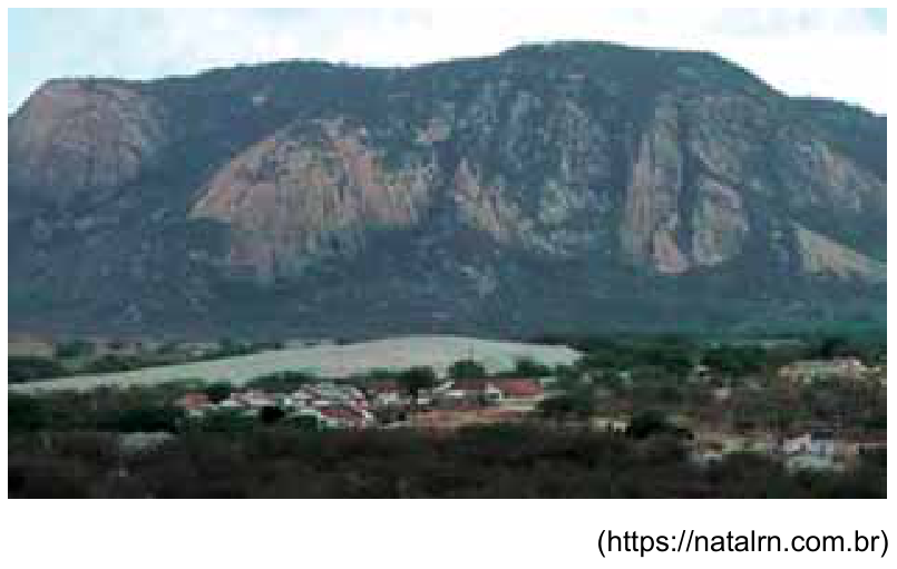Vista do Planalto da Borborema com relevo acentuado de formas convexas e rochosas ao fundo, vegetação e um pequeno povoamento em primeiro plano