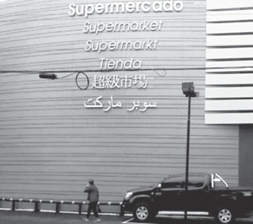 Fotografia da fachada de um supermercado com a palavra "supermercado" escrita em vários idiomas e sistemas de escrita: "Supermercado", "Supermarket", "Supermarkt", "Tienda", caracteres em chinês e escrita em árabe. Diante da fachada, vê-se uma calçada, um pedestre caminhando e uma caminhonete estacionada.