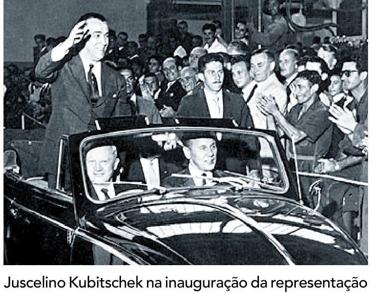 Fotografia em preto e branco de Juscelino Kubitschek em pé em um carro conversível, acenando para a multidão que o aplaude.