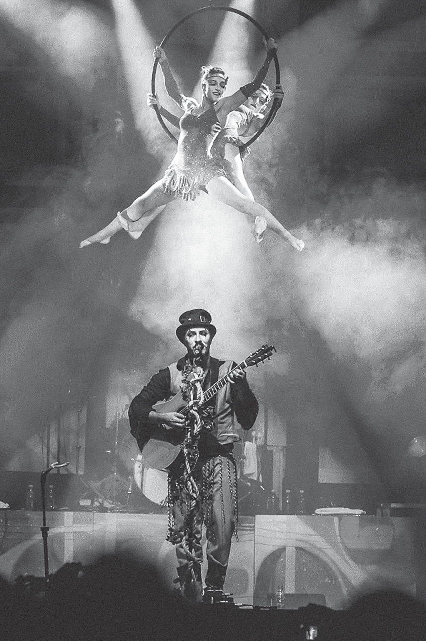 F1 Apresentação d'O Teatro Mágico: no alto, artistas circenses realizam acrobacias suspensas em uma lira acrobática sob iluminação cênica e fumaça; abaixo, um músico com maquiagem artística, cartola e figurino estilizado toca violão e canta no palco.