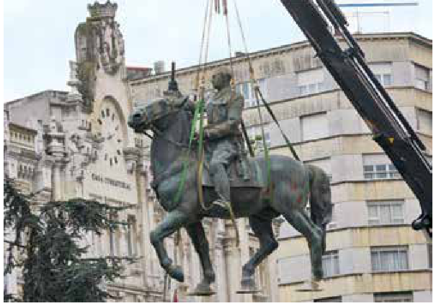 Fotografia da retirada da estátua equestre do General Francisco Franco, içada por cabos de sustentação presos a um guindaste, em frente a um edifício com relógio na fachada.