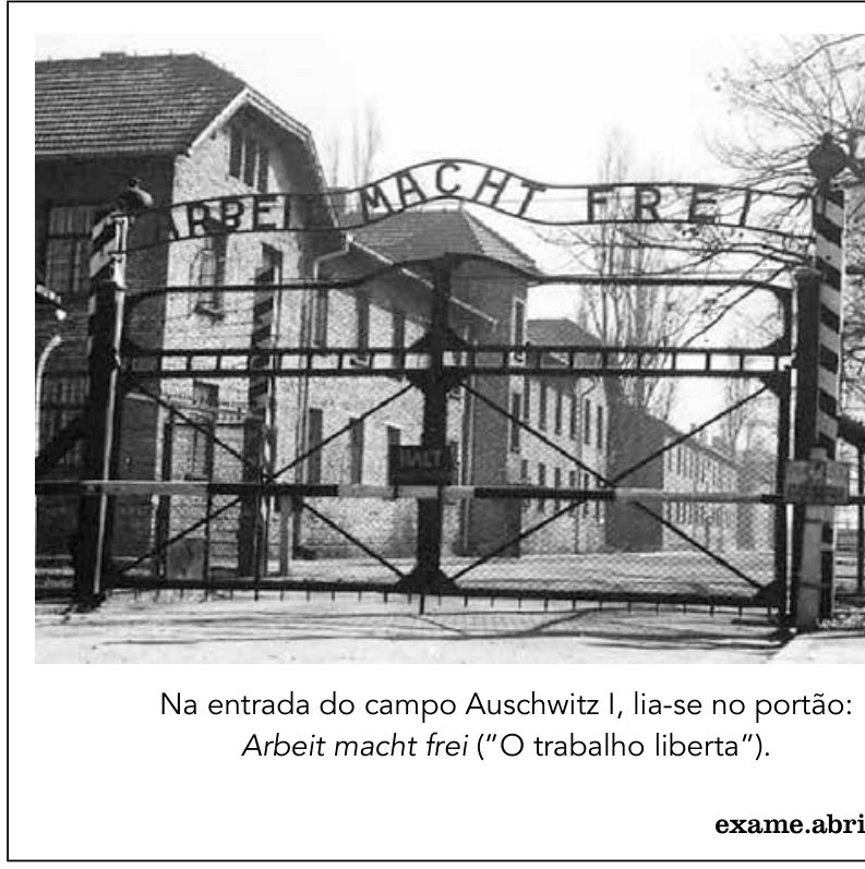 Fotografia em preto e branco do portão de entrada do campo de concentração de Auschwitz I, com a inscrição em ferro forjado "ARBEIT MACHT FREI". Abaixo da foto, lê-se a legenda: "Na entrada do campo Auschwitz I, lia-se no portão: Arbeit macht frei ('O trabalho liberta'). exame.abril"