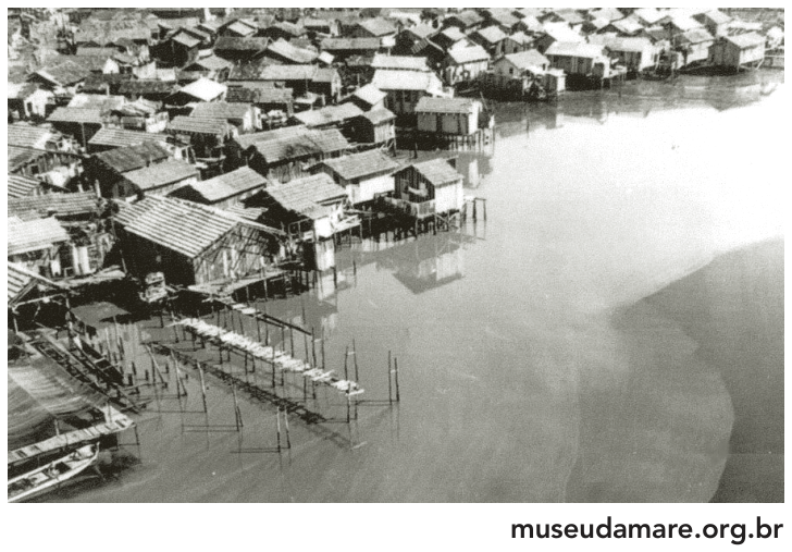 Fotografia em preto e branco da Baixa do Sapateiro, na Maré, com barracos de madeira sobre palafitas junto à água e passarelas de madeira ligando as moradias. Crédito: museudamare.org.br.