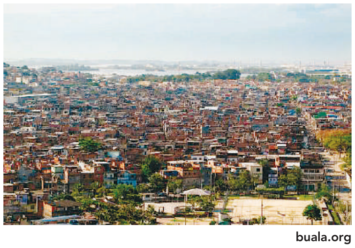 Fotografia panorâmica colorida da Maré, mostrando uma extensa ocupação urbana de construções de alvenaria muito próximas umas das outras, com algumas árvores e uma quadra na parte inferior. Crédito: buala.org.