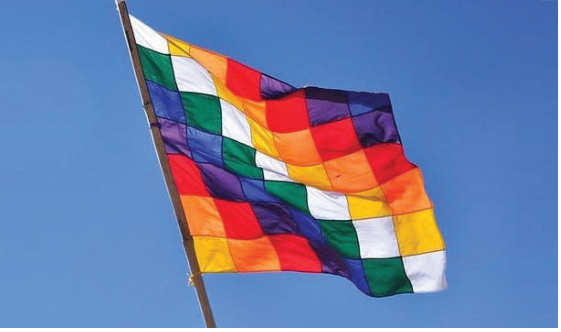 Fotografia da bandeira Wiphala, formada por uma grade de quadrados em sete cores dispostas em faixas diagonais, hasteada sob o céu azul.